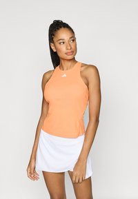 Orange athletic tank top with a racerback design and white logo, paired with a white, textured skort. Smooth fabric, fitted shape.