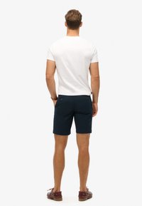 Mænds marineblå shorts lavet af bomuld, med baglommer med knapper, parret med en hvid tætsiddende t-shirt. Brune sko synlige.