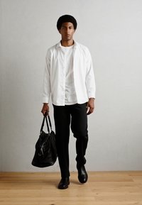 Sac en cuir noir, chemise blanche superposée sur un t-shirt blanc, pantalon noir et chaussures noires. Design lisse et minimaliste sur un sol en bois clair.