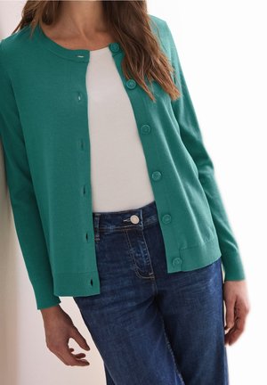 Mujer que lleva un cárdigan azul verdoso abotonado sobre una camiseta blanca y vaqueros de talle alto de color azul oscuro, de pie con postura relajada sobre fondo blanco.