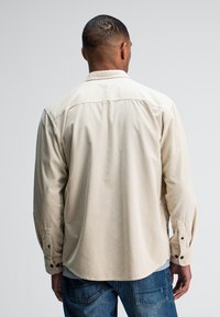 Beige overhemd met lange mouwen, een button-down kraag, twee knopen bij de manchetten en een rechte zoom, gecombineerd met blauwe denim jeans.