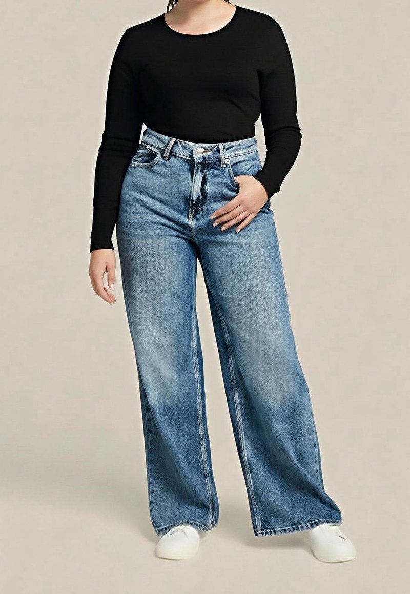 Personne portant un haut noir à manches longues, un jean bleu taille haute à jambes larges et des chaussures blanches, debout avec une main dans la poche.