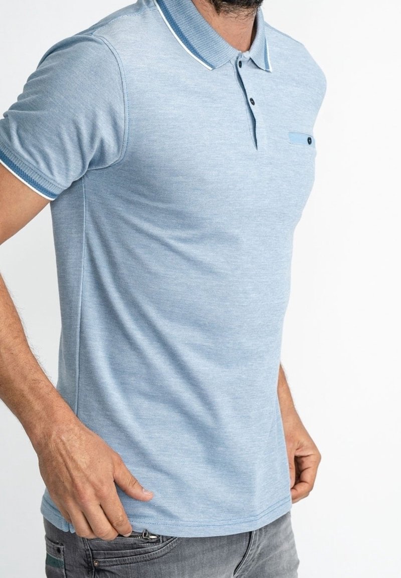 Hellblaues Poloshirt mit strukturierter Webart, kontrastierendem dunkelblauem Kragen und mit Knöpfen versehenem Brusttasche, kombiniert mit grauen Jeans.