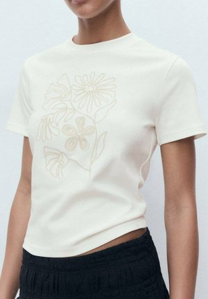 Modèle portant un t-shirt crème à manches courtes avec broderie florale, associé à un short noir texturé, sur fond gris clair.