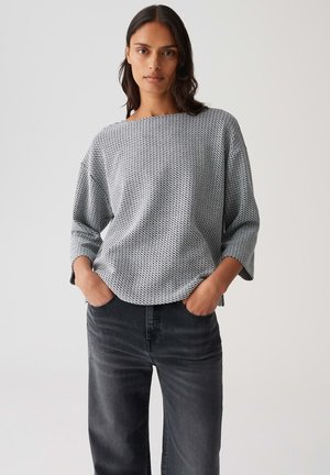 Femme aux cheveux noirs portant un haut gris ample à motifs et un jean noir, se tenant avec les mains dans les poches devant un fond clair.