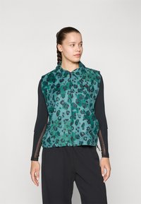 Gilet à imprimé léopard vert et noir, avec un col et une fermeture éclair à l'avant, fabriqué dans un tissu lisse, associé à un haut à manches longues noir.