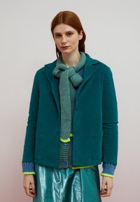 Teal farvet fløjlsblazer med skræddersyet design, der har en hakket krave, to forlommer og et tekstureret grønt strikket tørklæde med gule accenter.