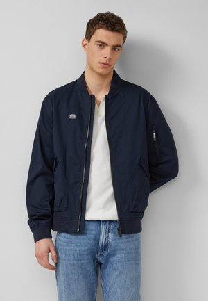 Jeune homme portant une veste bomber bleu marine sur une chemise blanche et un jean bleu clair, debout devant un fond gris clair uni.