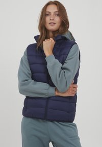 Marineblauwe gewatteerde vest met hoge kraag en ritssluiting aan de voorkant, gedragen over een lichtblauwe sweatshirt, met ribgebreide manchetten en onderrand.