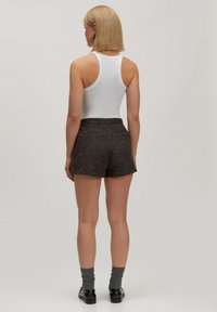 Vrouw met een blonde bob die een witte mouwloze top, gestructureerde donkere shorts, grijze sokken en zwarte schoenen draagt, staand met haar rug naar ons gekeerd.