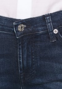 Jeans de mezclilla azul oscuro con cierre frontal de botón, costuras en contraste y una textura suave. La cinturilla tiene acentos de metal.