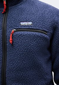 Αντιανεμικό μπουφάν από fleece σε ναυτικό μπλε χρώμα με υφασμάτινη υφή, μαύρο φερμουάρ με κόκκινο τράβηγμα και λογότυπο Patagonia πάνω από μια τσέπη στο στήθος με φερμουάρ.
