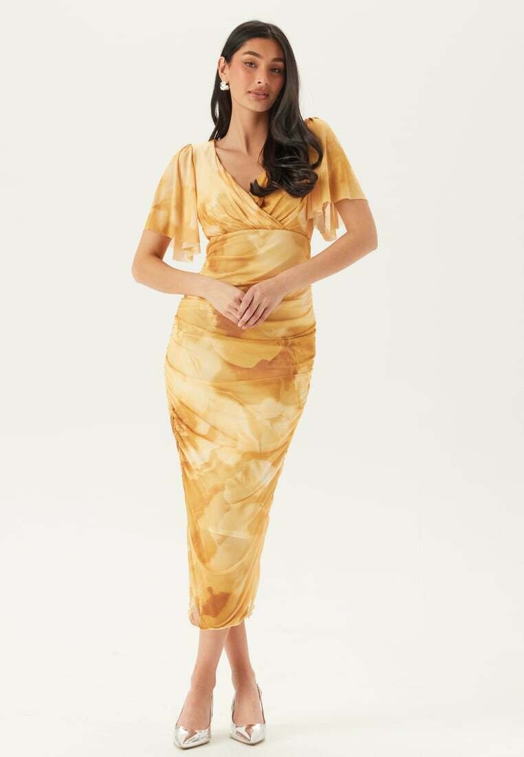 Donna con lunghi capelli scuri che indossa un abito avvolgente giallo a fiori e tacchi argento, in piedi con le mani intrecciate davanti a sé.