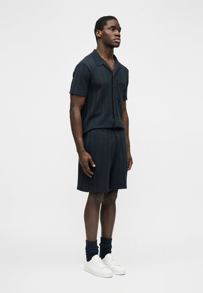 Jeune homme debout face à gauche portant une chemise à manches courtes texturée sombre, un short assorti, des chaussettes foncées et des baskets blanches sur un fond uni.