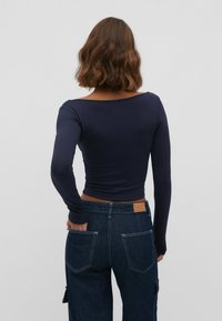 Haut à manches longues bleu marine avec un large décolleté, associé à un pantalon en denim foncé taille haute avec deux poches arrière et une étiquette en cuir marron clair.