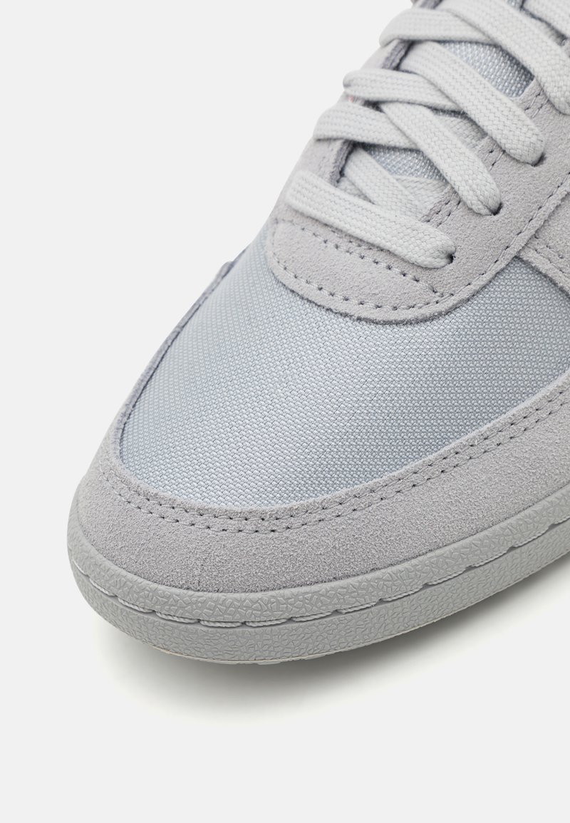Helle graue Sneaker aus einer Kombination von glattem Synthetikmaterial und strukturiertem Wildleder, mit flachen Schnürsenkeln und einer Gummisohle.