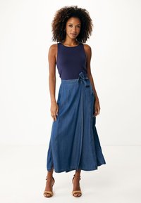 Džínová maxi sukně s předním zavazováním, která má rozšířený střih, vybledle modrou barvu a jemné šití. Kombinováno s tmavým tílkem a podpatky.