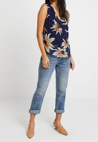 Top senza maniche blu navy con motivo floreale in crema e arancione, abbinato a jeans azzurri arrotolati e scarpe con tacco marroni chiari.