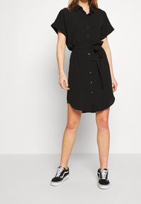 Vestido corto negro de manga corta con botones en el frente y cinturón. Presenta un dobladillo redondeado y tela suave. Combinado con zapatillas negras.