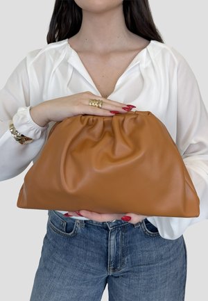 Donna con camicetta bianca e jeans tiene una grande pochette in pelle marrone chiaro, con unghie rosse, anello d'oro e braccialetto, su sfondo grigio.