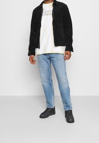Black corduroy jacket with a zip closure, worn over a white tie-dye t-shirt, light blue jeans, and black sneakers. Simple design, no patterns.