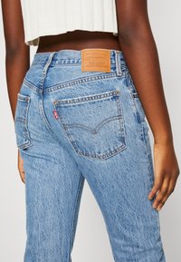 Jeans de denim azul claro con cintura alta, que presentan un parche de cuero, costuras naranjas y bolsillos traseros con un diseño distintivo.