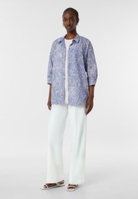 Light blue floral-printed shirt jacket with a relaxed fit, three-quarter sleeves, button closure, worn over a white top and paired with white wide-leg pants.