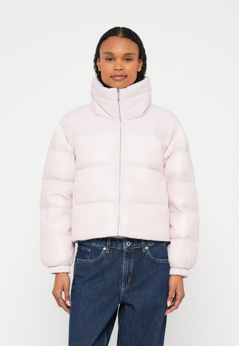 Femeie purtând o geacă matlasată roz pal cu guler înalt și fermoar, asortată cu blugi largi de culoare albastru închis, stând în fața unui fundal alb.