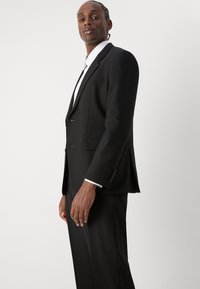 Schwarze Sakkjacke mit Revers, zwei Fronttaschen und einem Ein-Knopf-Verschluss. Kombiniert mit einem weißen Hemd, elegantes Design.