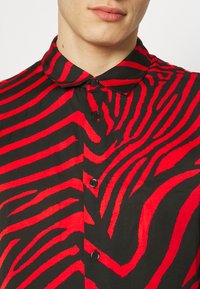 Maglietta nera con audace motivo a strisce zebra rosse e nere, con collo a camicia e bottoni sulla parte anteriore. Tessuto dalla texture liscia.