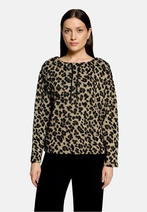 Kvinde med glat mørkt hår iført en langærmet beige og sort leopardprintbluse og sorte bukser mod en ensfarvet baggrund.