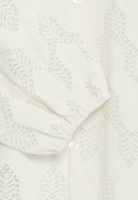 White blouse with a textured, lace-like pattern featuring diamond shapes and leaf motifs. Includes subtle button closure at the cuff.
