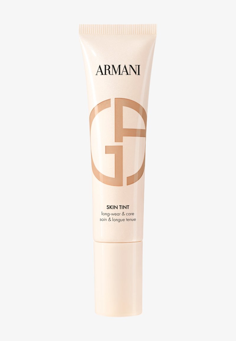 Kremfarget tube med en blank finish, med en fremtredende beige logo og svart tekst. Etiketten angir "Skin Tint" og "langvarig og pleie."