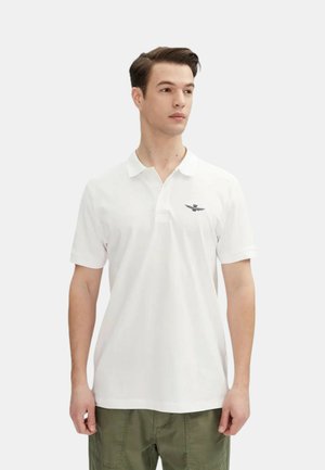 Polo bianco realizzato in cotone, con maniche corte, un'apertura con due bottoni e un piccolo emblema ricamato sul lato sinistro del petto.