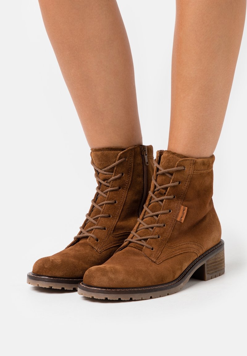 Bottines marron en daim à lacets avec fermeture éclair latérale et talons carrés portées sur jambes nues.