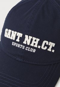 Navy blue cap made of textured fabric featuring white embroidered text: "GANT NH. CT. SPORTS CLUB." Includes stitched accents around the brim.