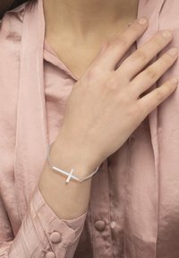 Sølvarmbånd med minimalistisk korsdesign, med en glatt, polert overflate og en sammenkoblet kjede. Båret på en hånd med en myk rosa erme.