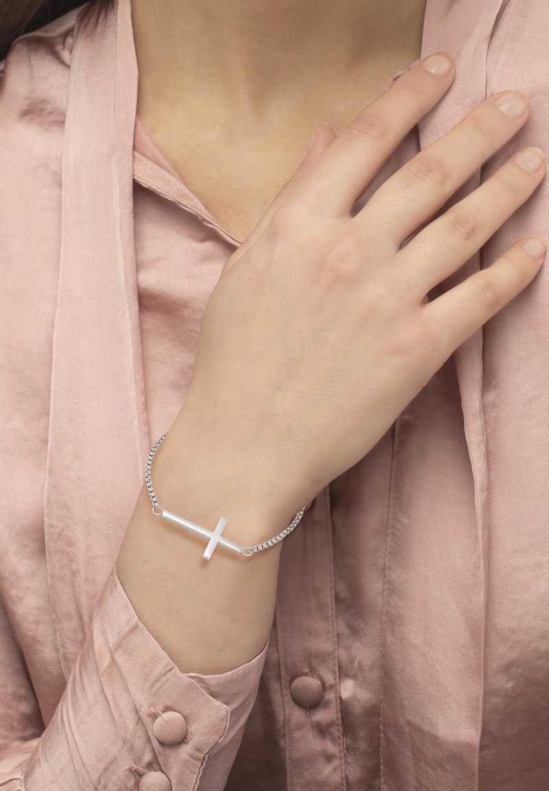 Sølvarmbånd med minimalistisk korsdesign, med en glatt, polert overflate og en sammenkoblet kjede. Båret på en hånd med en myk rosa erme.
