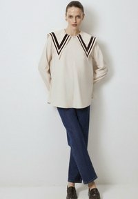 Beige blouse met contrasterende bruine fluwelen chevronkraag, lange mouwen en een rechte snit. Gecombineerd met donkerblauwe rechte jeans.