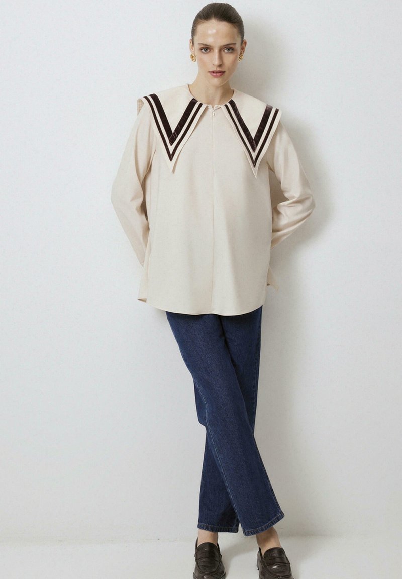 Beige blouse met contrasterende bruine fluwelen chevronkraag, lange mouwen en een rechte snit. Gecombineerd met donkerblauwe rechte jeans.