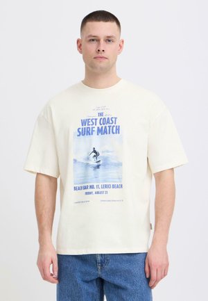 Homme portant un T-shirt crème à manches courtes avec un imprimé d'événement de surf bleu, associé à un jean bleu, debout devant un fond blanc uni.
