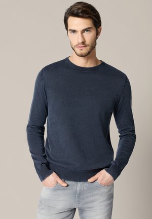 Mężczyzna z ciemnymi włosami i brodą, ubrany w granatową koszulę z długim rękawem oraz jasnoszare jeansy, stojący na tle gładkiego beżowego tła.