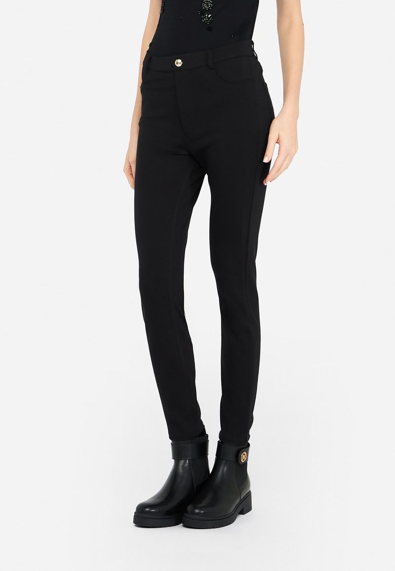 Pantalon noir slim avec détail de bouton doré, associé à des bottines noires avec un emblème rond doré sur le côté.
