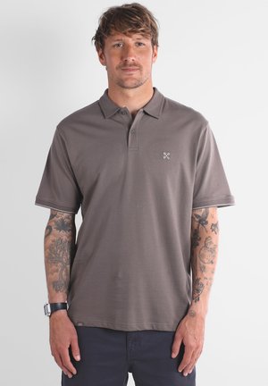 Homme aux cheveux bruns portant un polo gris et un pantalon sombre, affichant des tatouages sur les deux avant-bras et une montre au poignet gauche.