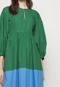 Robe verte avec un col rond et un détail en forme de trou de serrure. Présente une taille froncée et des manches volumineuses. La partie inférieure est bleue. Texture en coton.