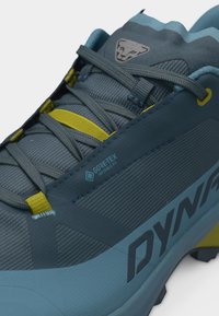 Κοντινό πλάνο ενός πεζοπορικού παπουτσιού Dynafit σε μπλε και κίτρινες αποχρώσεις, με υλικό Gore-Tex Invisible Fit και γκρι κορδόνια.