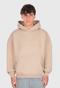 Un jeune homme aux cheveux courts et bouclés porte un sweat à capuche beige avec une poche sur le devant et un texte en relief "DROPSTYLE", associé à un jean bleu clair.