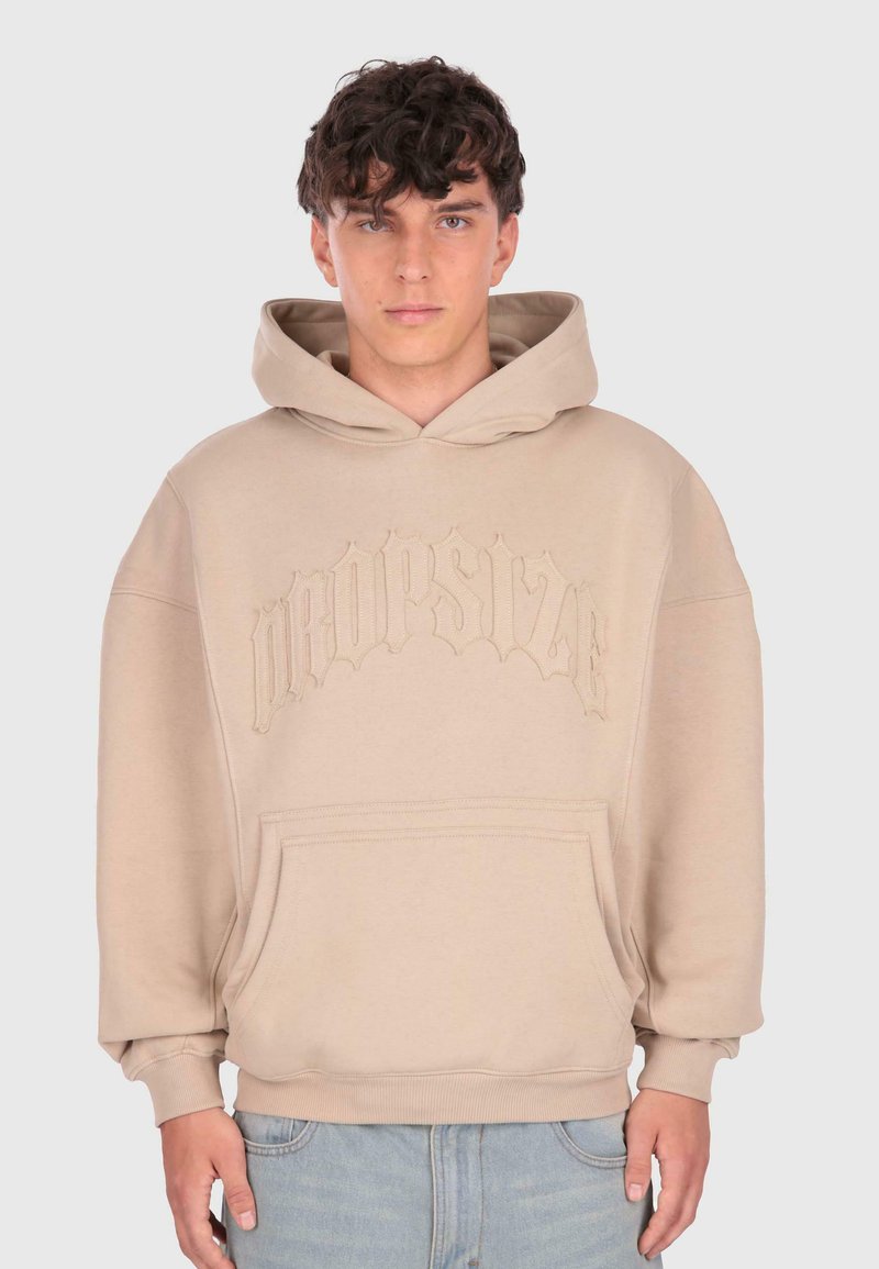 Un jeune homme aux cheveux courts et bouclés porte un sweat à capuche beige avec une poche sur le devant et un texte en relief "DROPSTYLE", associé à un jean bleu clair.