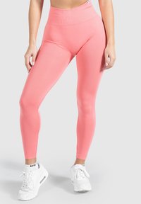 Leggings corallo a vita alta realizzati in un tessuto elasticizzato e texturizzato. Abbinati a scarpe da ginnastica bianche con lacci prominenti e logo visibile.