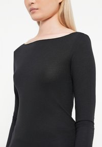Donna con lunghi capelli biondi che indossa un top nero a maniche lunghe aderente con un ampio scollo su uno sfondo bianco.